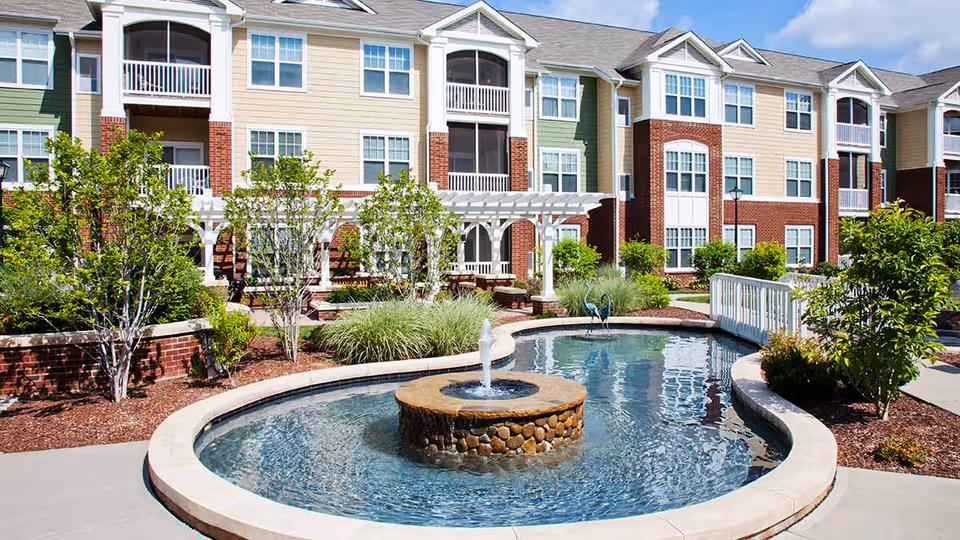 Outdoor courtyard area of a senior living facility featuring a decorative water fountain in a pond, surrounded by landscaped greenery, a white pergola, and a multi-story building with balconies and large windows in the background.