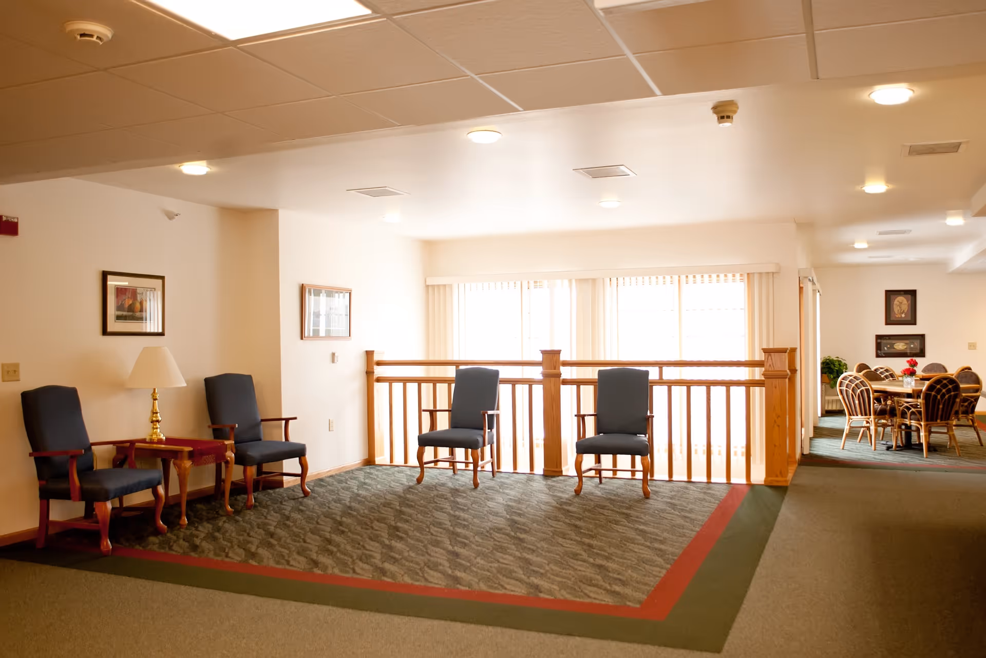 A well-lit interior common area with four wooden chairs with blue cushions arranged around a small wooden table with a lamp. The area has carpeted flooring with a green and red border. In the background, there is a dining area with a round table and several chairs. The walls are decorated with framed pictures.