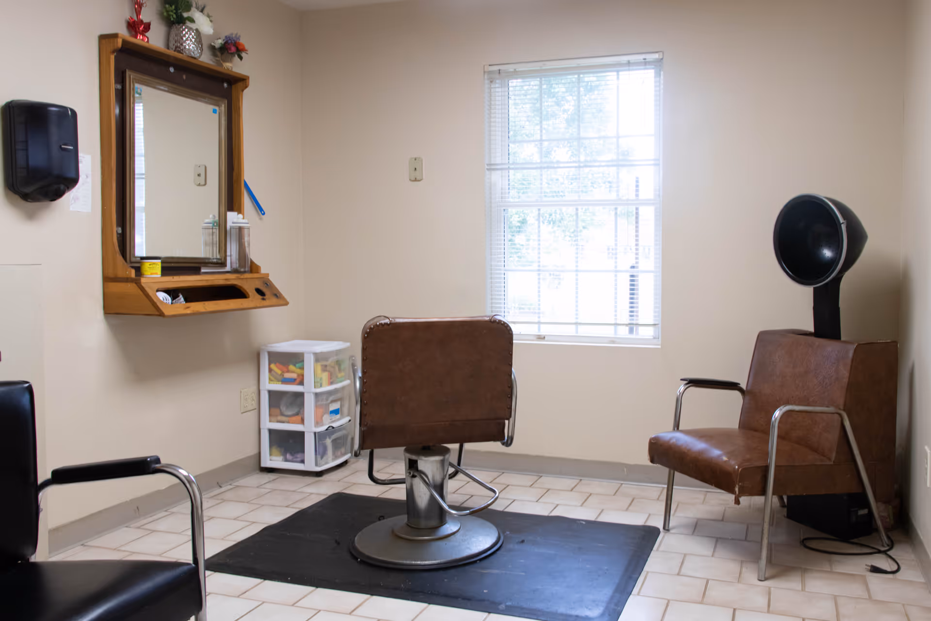 A small salon room with a brown salon chair in front of a wall-mounted mirror and shelf. To the right, there is a brown chair with a black hair dryer hood attached. A window with blinds lets in natural light, and a small plastic drawer unit with various items is placed near the corner.