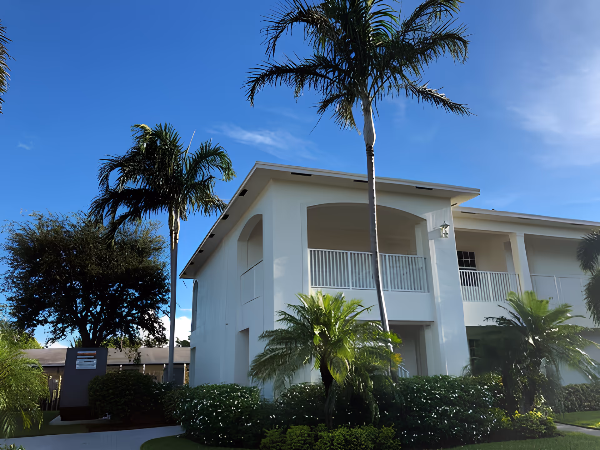 Exterior view of a two-story white building with a balcony, surrounded by palm trees and lush green bushes under a clear blue sky.