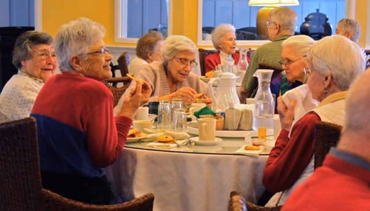 A group of elderly people sitting around a round dining table in a well-lit room, enjoying snacks and drinks while engaging in conversation.