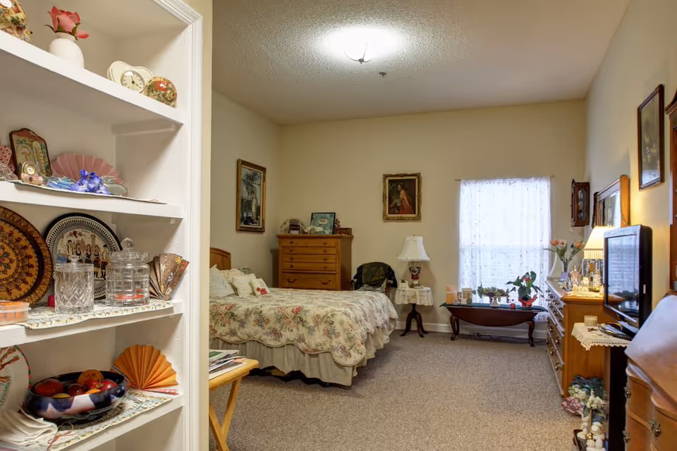 Comfortable bedroom in a senior living suite with a floral bed, wooden dressers, display shelves of knickknacks, and a TV.