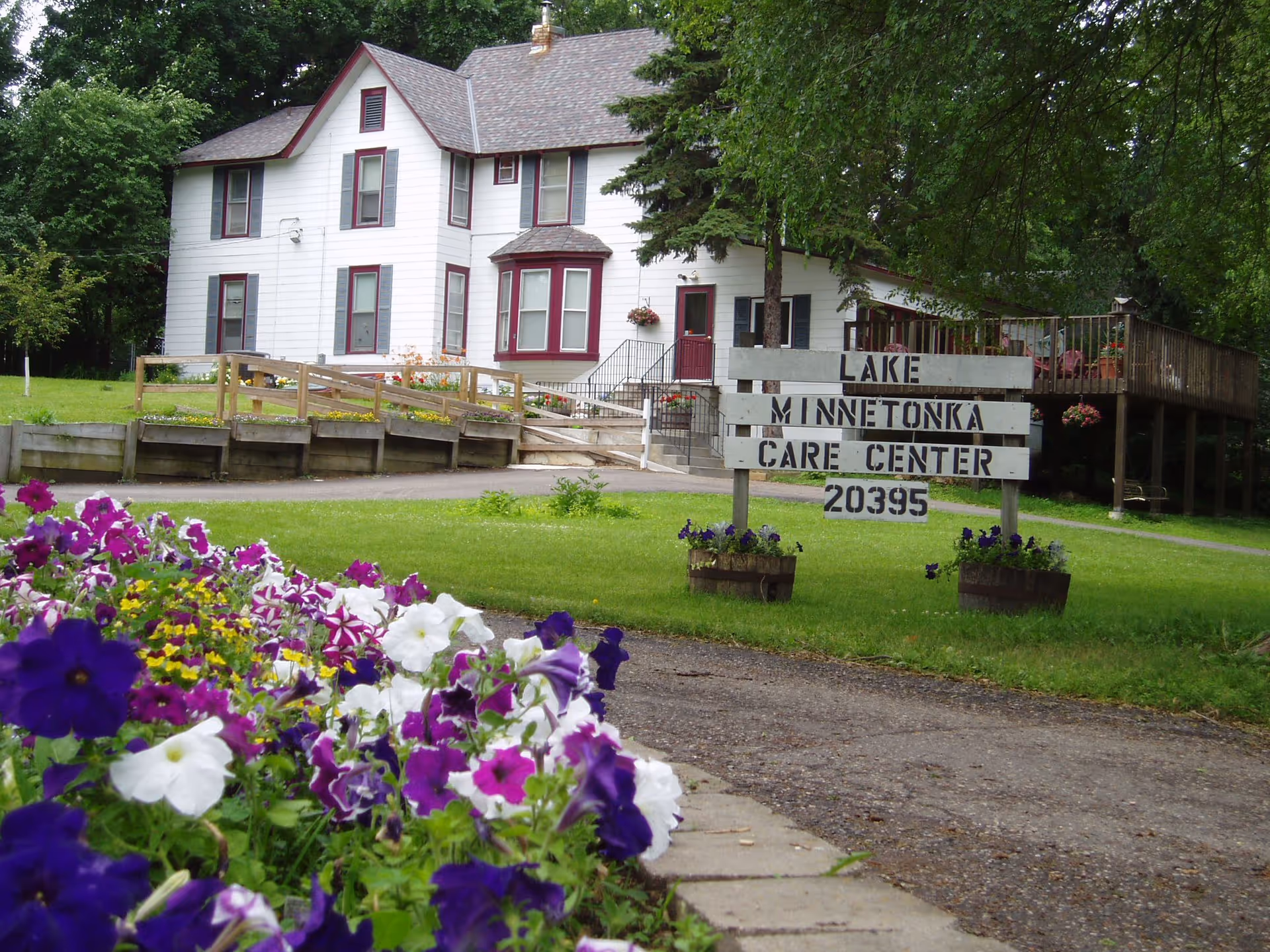 A white two-story building with maroon window frames and a wooden ramp leading to the entrance. In front of the building is a wooden sign that reads 'Lake Minnetonka Care Center 20395'. There are colorful flowers in the foreground and green trees surrounding the area.