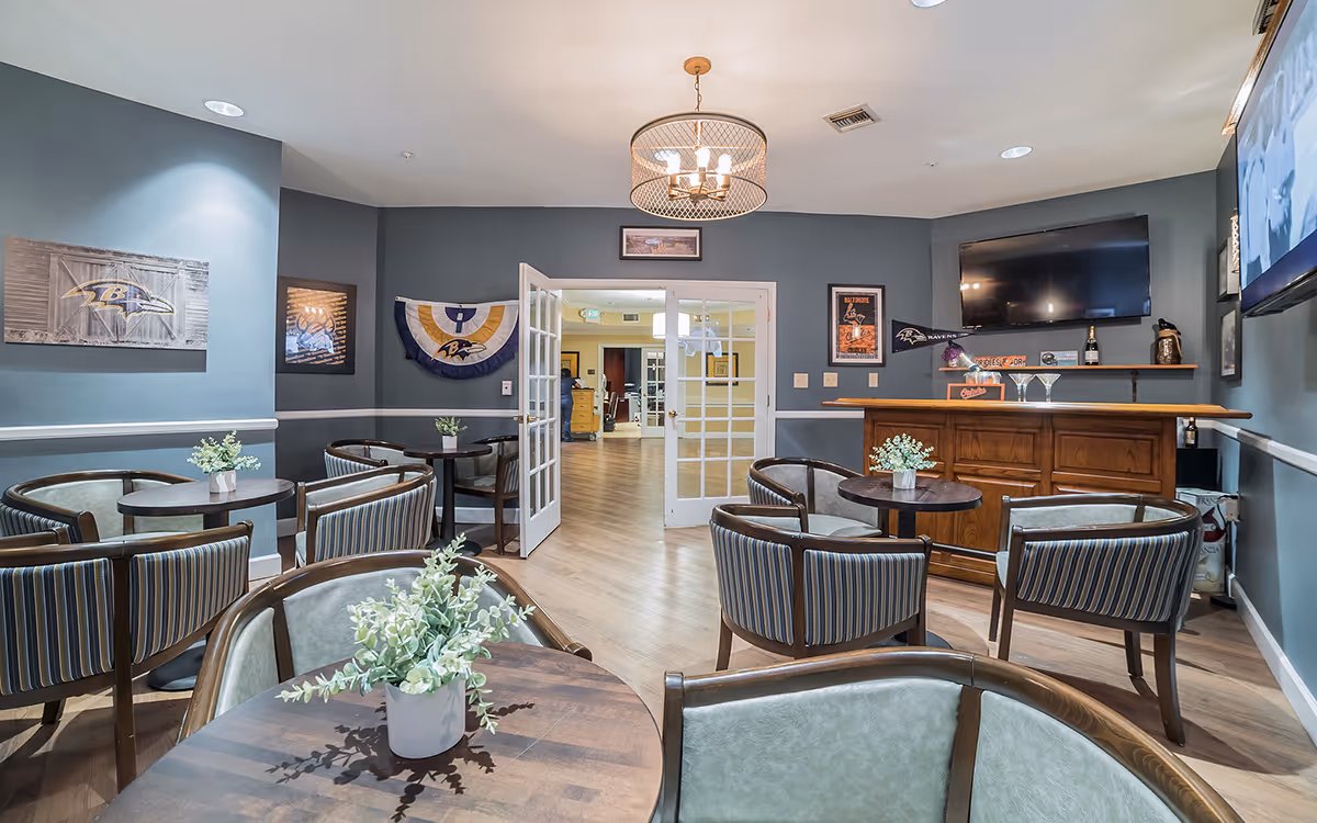 A cozy common area with several round tables and cushioned chairs arranged around them. Each table has a small potted plant centerpiece. The walls are painted gray with white trim and decorated with Baltimore Ravens sports memorabilia. A wooden bar with a few glasses and bottles is on the right side, beneath a mounted flat-screen TV. Double glass doors lead to another room with light-colored walls and wooden flooring.
