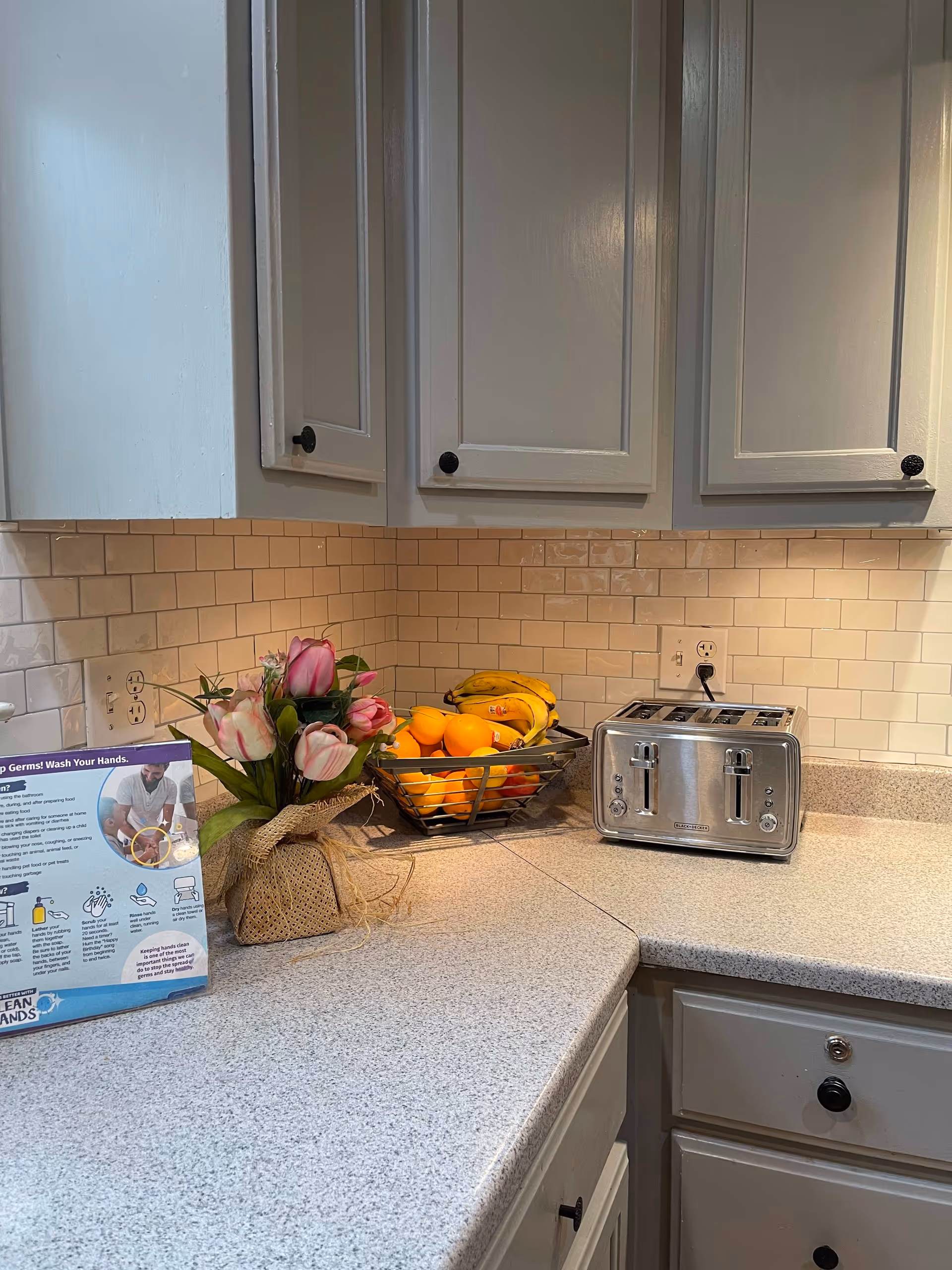 Kitchen countertop with white cabinets and subway tile backsplash holding a toaster, a fruit basket, and a vase of flowers.