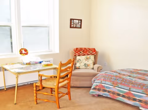 Simple bedroom with a patterned-bed, an upholstered armchair with pillows, a small table by a window and a wooden chair.