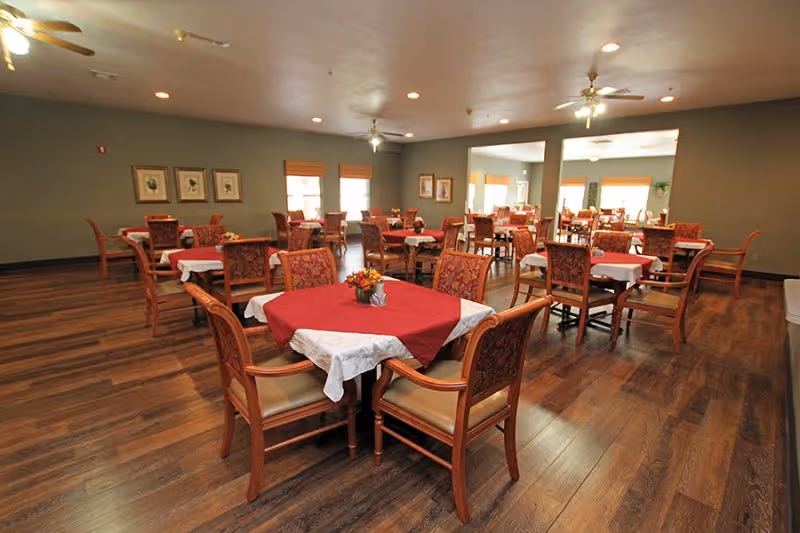 Spacious dining room with multiple tables set with red and white tablecloths and wooden chairs on hardwood floors.