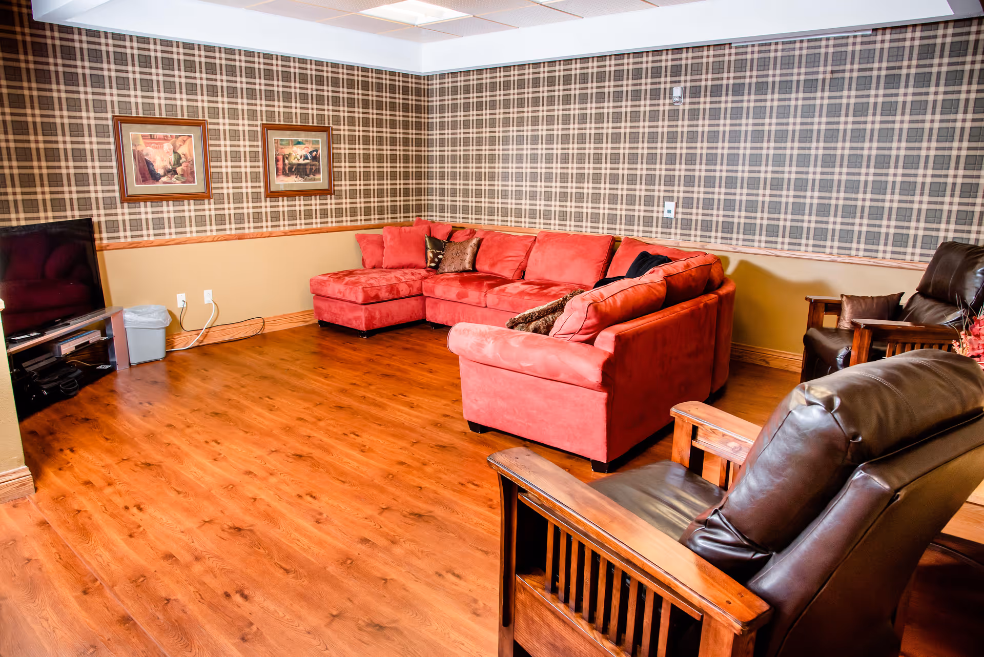 Cozy senior living lounge with a red sectional sofa, leather armchairs, wood floors, TV, and plaid wallpaper.