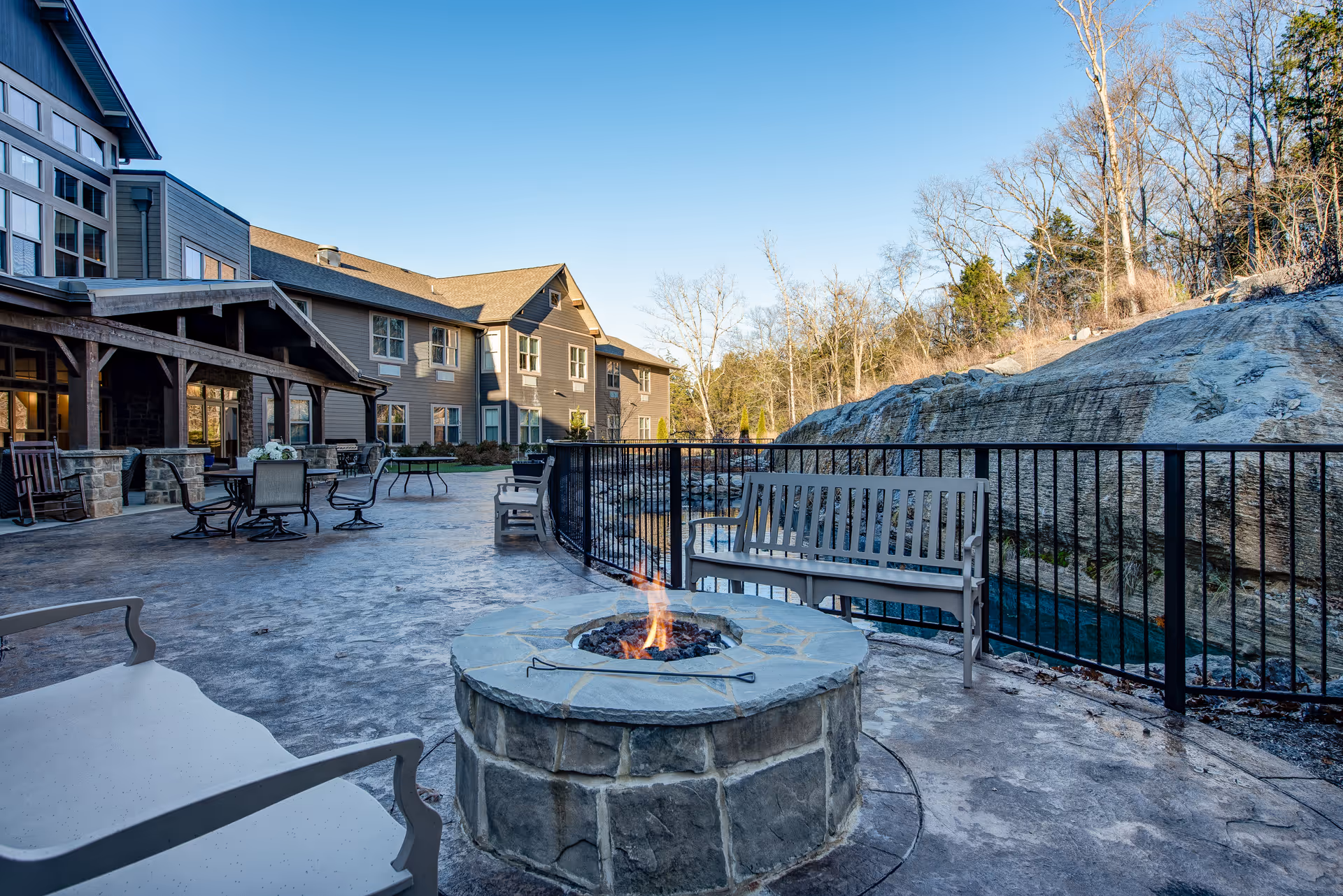 Outdoor patio area at Northshore Heights featuring a stone fire pit with a small flame, surrounded by benches and chairs. The patio overlooks a rocky landscape with trees in the background under a clear blue sky. The building exterior with large windows and a covered seating area is visible on the left side.