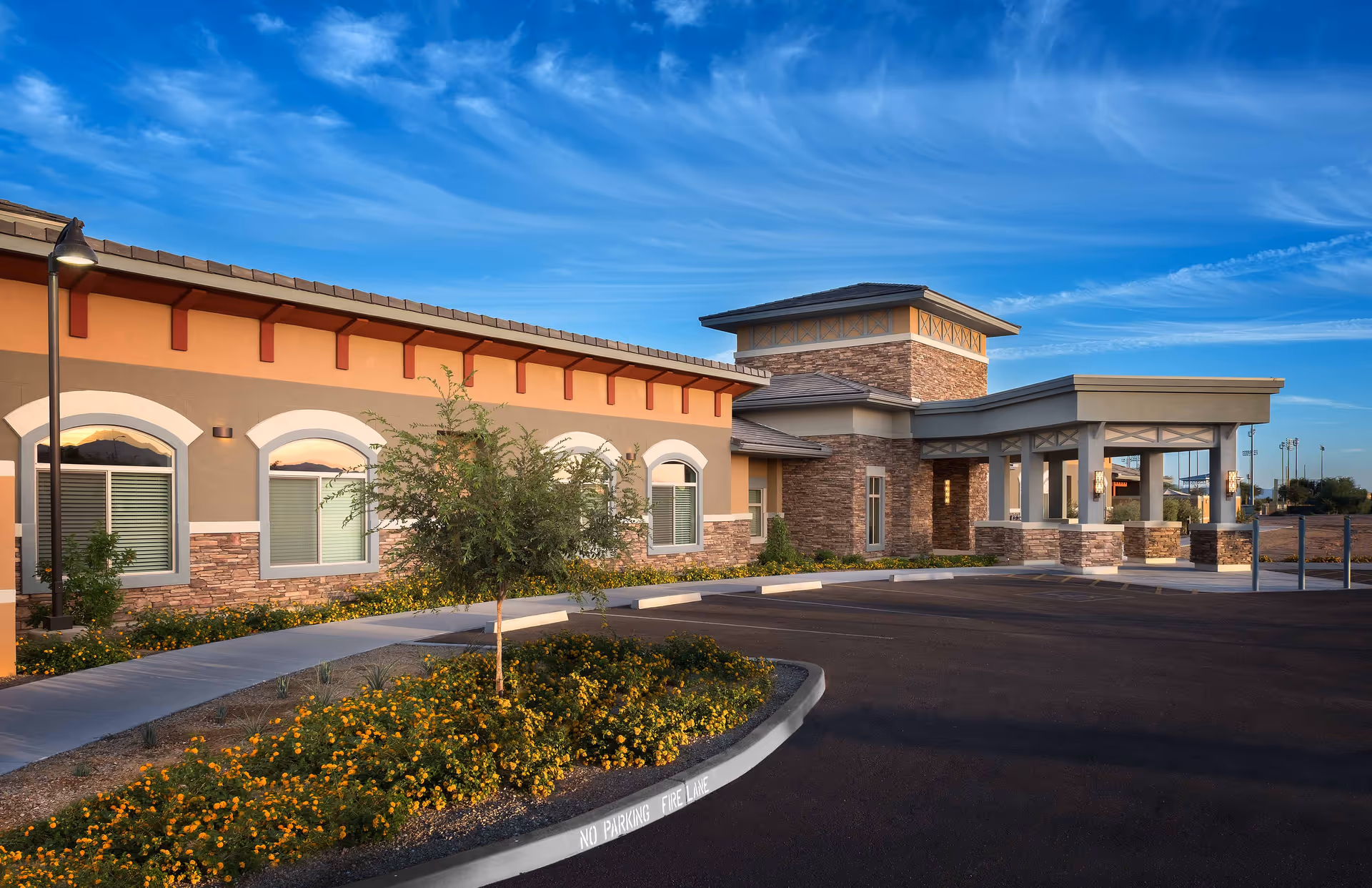 Exterior view of Mosaic Gardens Memory Care at Surprise facility showing a single-story building with stone and stucco facade, arched windows, a covered entrance, landscaped flower beds with yellow flowers, a small tree, and a clear blue sky with wispy clouds.