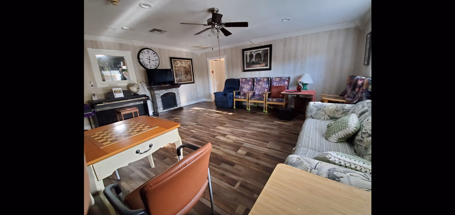 A senior living common room with hardwood floors, a sofa and several armchairs, a ceiling fan, TV over a fireplace, and a small table with a chessboard.