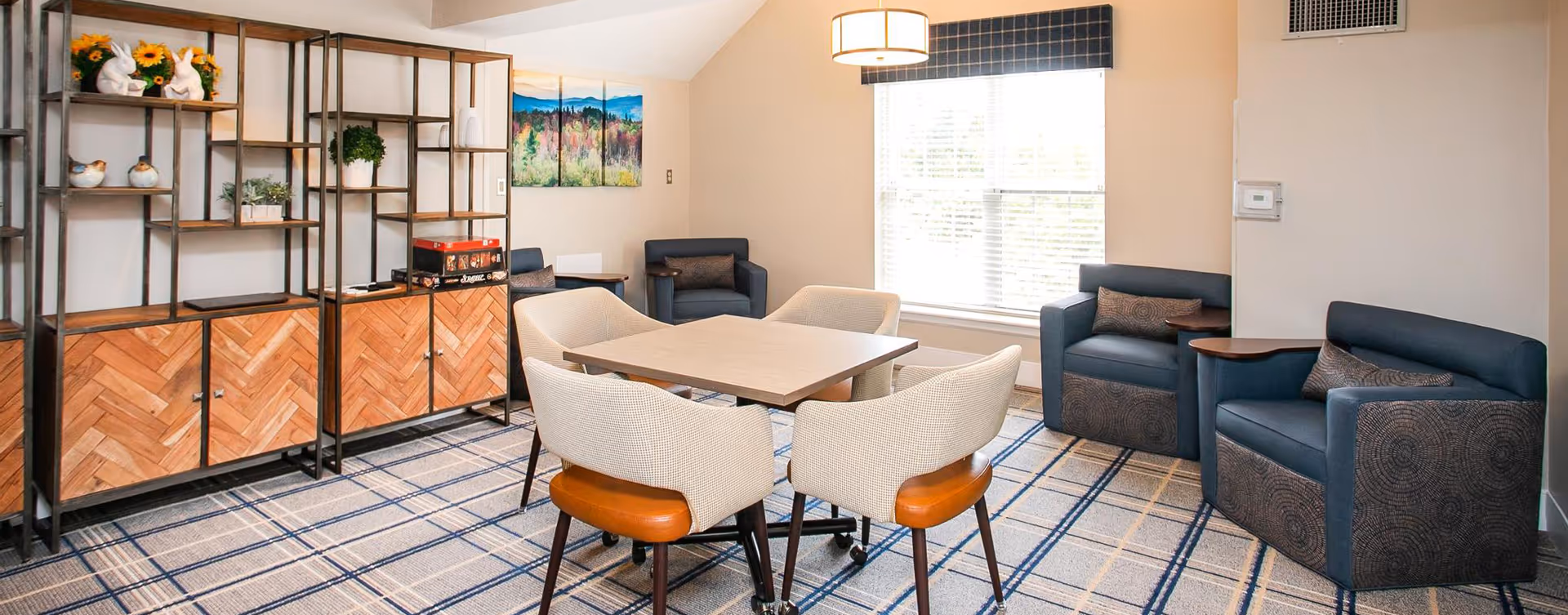 Well-lit communal sitting area with a square table surrounded by four chairs, upholstered armchairs by a window, and wooden shelving along the wall.