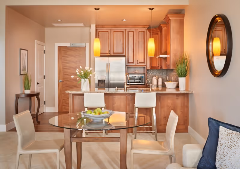 A modern kitchen and dining area with wooden cabinets, a stainless steel refrigerator, and a granite countertop island with two white bar stools. A round glass dining table with four beige chairs is in the foreground, with a bowl of green apples on the table. The room is warmly lit with two pendant lights hanging over the island, a round mirror on the wall, and decorative plants placed around the space.