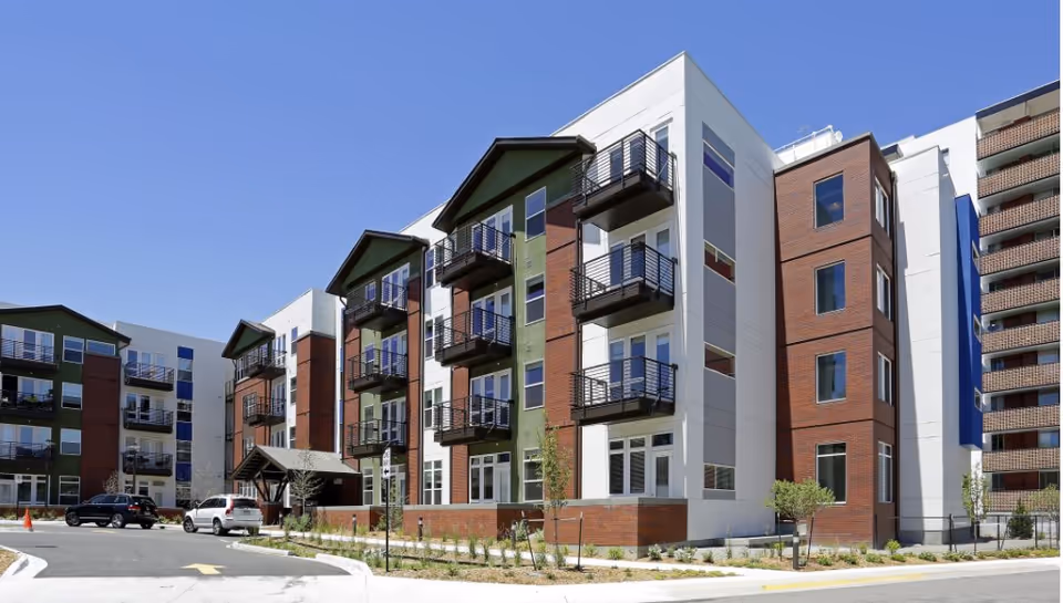 Exterior view of a modern multi-story residential building with balconies, large windows, and a combination of white, green, and brown facade under a clear blue sky. There are parked cars and a driveway in front of the building.
