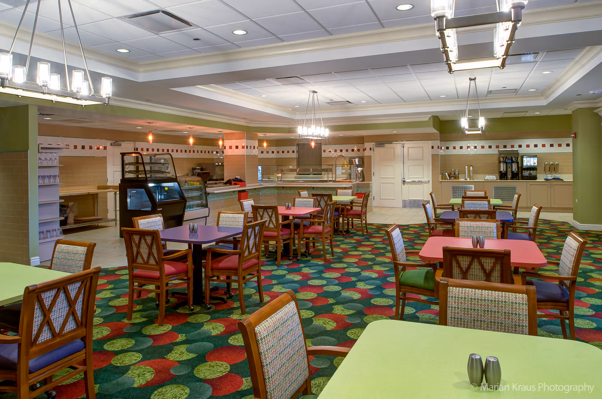 A brightly lit dining room with colorful tables and chairs arranged on a patterned carpet. The room features a serving counter with food display cases and beverage dispensers along the back wall. Modern light fixtures hang from the ceiling, and the walls are painted in light green and beige tones.