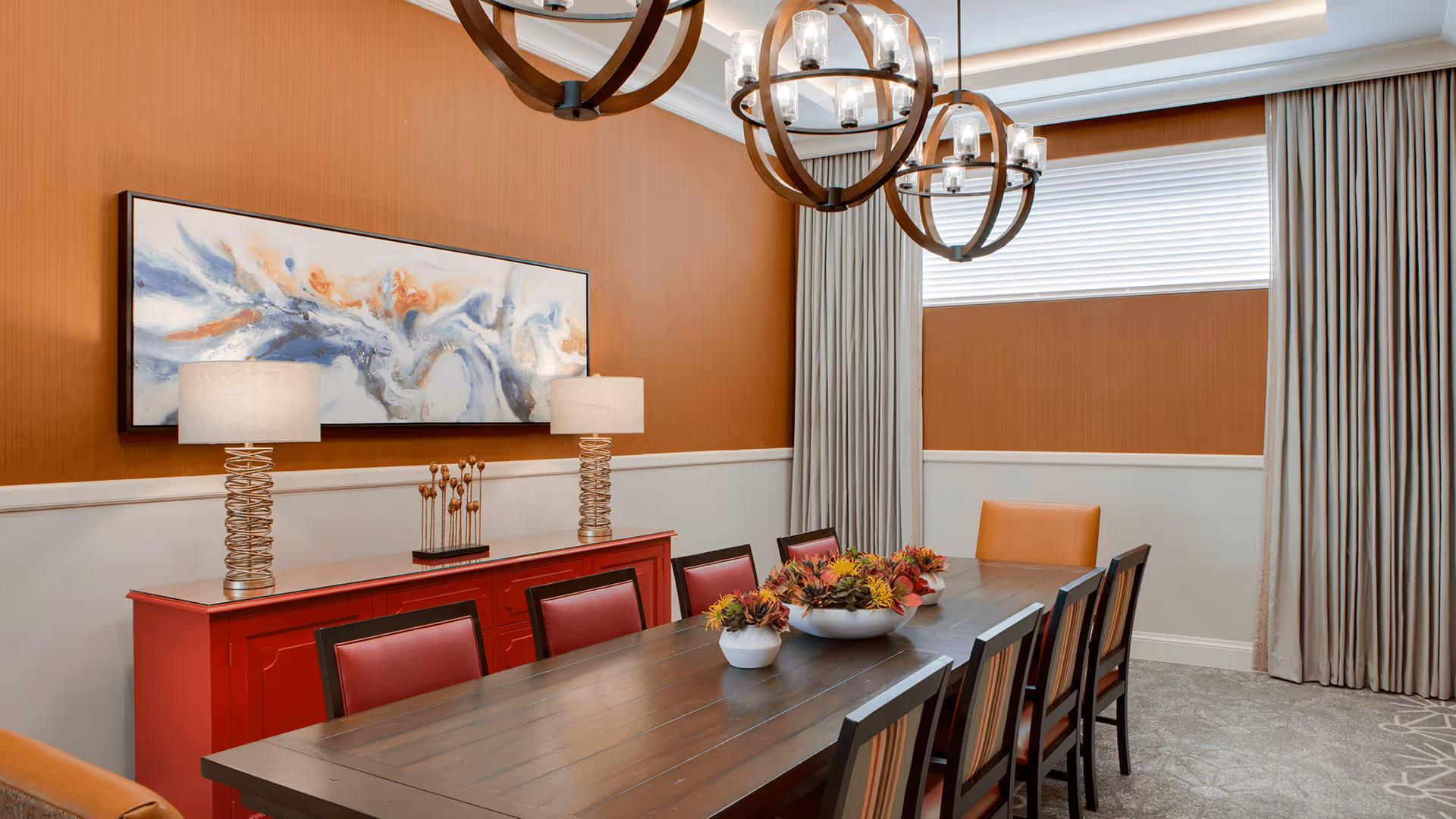 A dining room with a long wooden table surrounded by eight chairs, a red sideboard with two modern lamps and decorative items, an abstract painting on the orange wall, large windows with beige curtains, and two round chandeliers hanging from the ceiling.