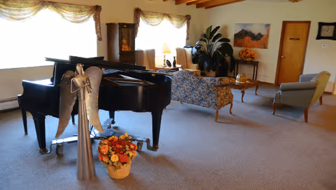 A spacious common lounge with a black grand piano, upholstered sofas and chairs, plants, and floral arrangements.