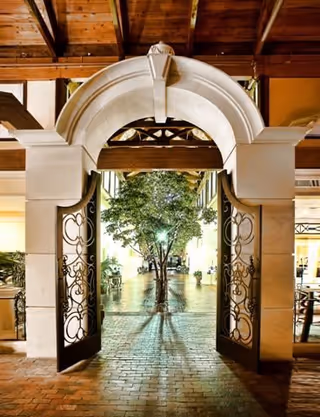 An open ornate wrought iron gate set in a large stone archway leading to a courtyard with a tree in the center, illuminated by soft lighting at night. The area has a warm, inviting ambiance with wooden beams overhead and brick flooring.