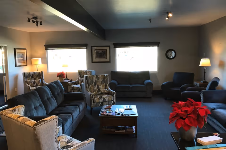 A cozy senior living facility common area with multiple upholstered sofas and armchairs arranged around a wooden coffee table. The room has two large windows with blinds, framed artwork on the walls, and several lamps providing warm lighting. A red poinsettia plant is placed on a side table in the foreground.