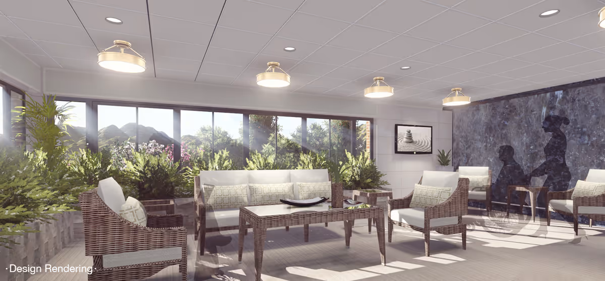 A bright and spacious indoor lounge area with large windows showing greenery outside. The room features wicker furniture with cushions, including chairs and a sofa around a coffee table. There are several ceiling lights, plants, and a wall decoration with silhouettes of a person pushing another in a wheelchair.