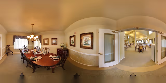 A panoramic view of a dining room in a senior living facility named Marbella Chico. The room features a long wooden dining table set with plates, glasses, and napkins, surrounded by elegant wooden chairs. The walls are decorated with framed artwork and a sideboard with a floral arrangement. Double glass doors open into a larger room with more dining tables and chairs, illuminated by ceiling lights and natural light from windows.