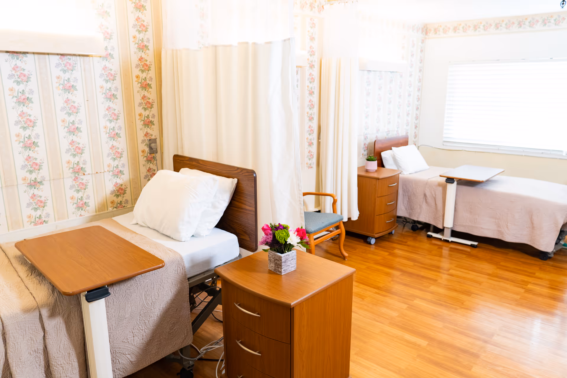 A bright and clean room in a senior living facility with two hospital-style beds separated by a privacy curtain. Each bed has a wooden overbed table, white pillows, and beige blankets. The room features floral wallpaper, wooden flooring, a small wooden nightstand with a flower pot, and a chair with wooden arms. A large window with blinds allows natural light to fill the space.