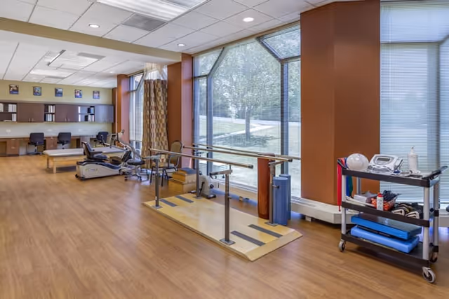 Interior rehabilitation gym with parallel bars, exercise equipment, and large windows.