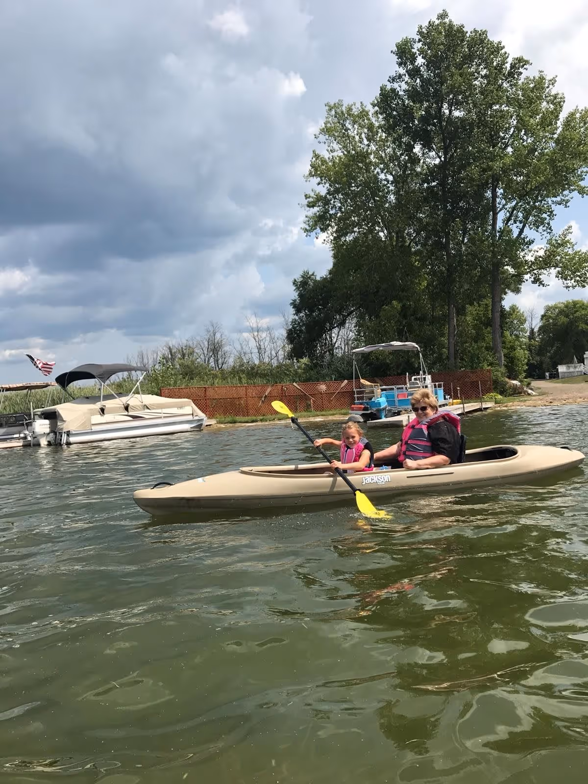 An older woman and a young girl wearing pink life jackets are kayaking together on a lake near a dock with boats and trees in the background under a cloudy sky.