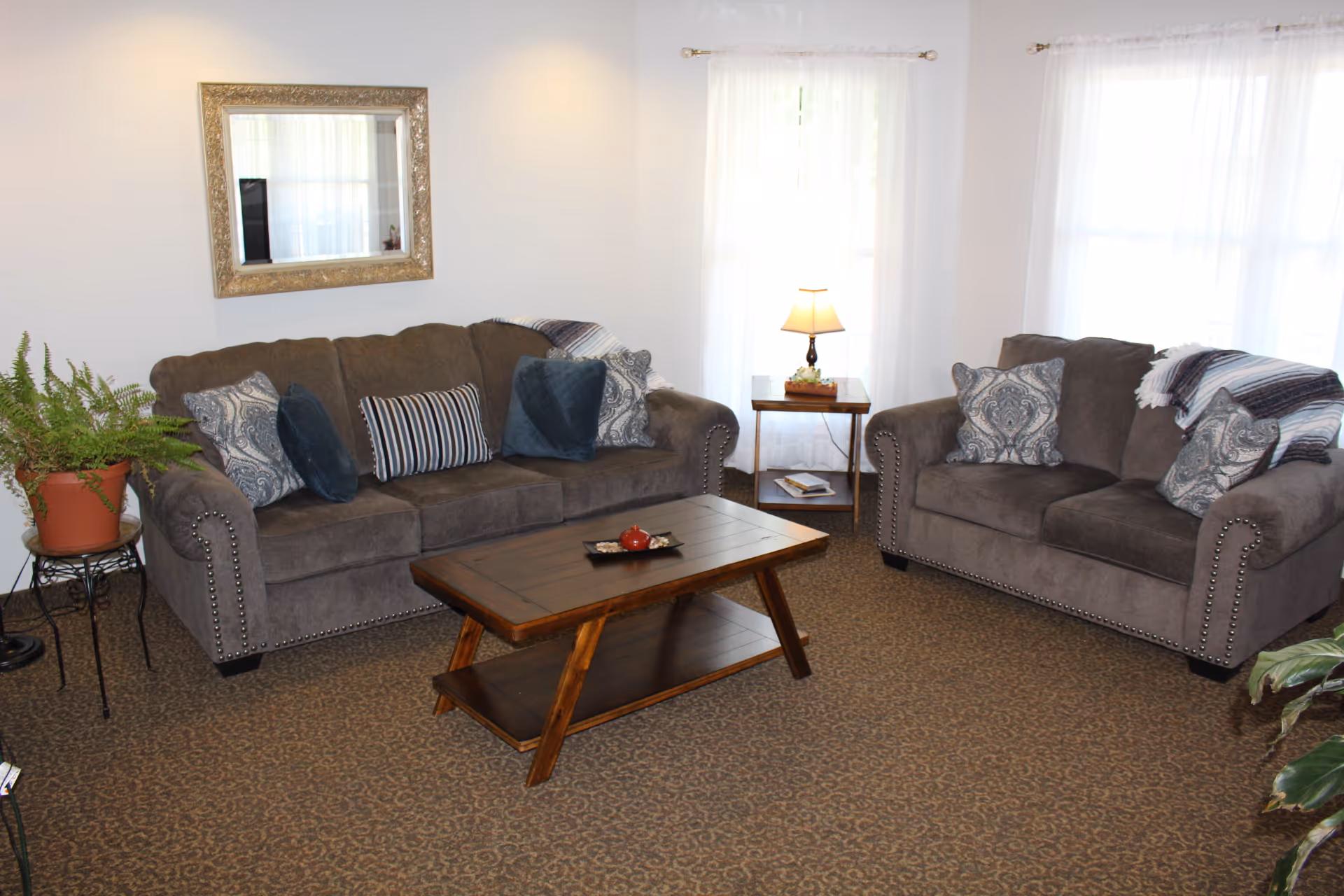 A cozy living room with two gray upholstered sofas adorned with patterned and solid pillows. A wooden coffee table sits in the center on a patterned carpet. There is a small side table with a lamp between two windows with sheer white curtains. A potted plant is on a stand to the left, and a large decorative mirror hangs on the wall above one sofa.