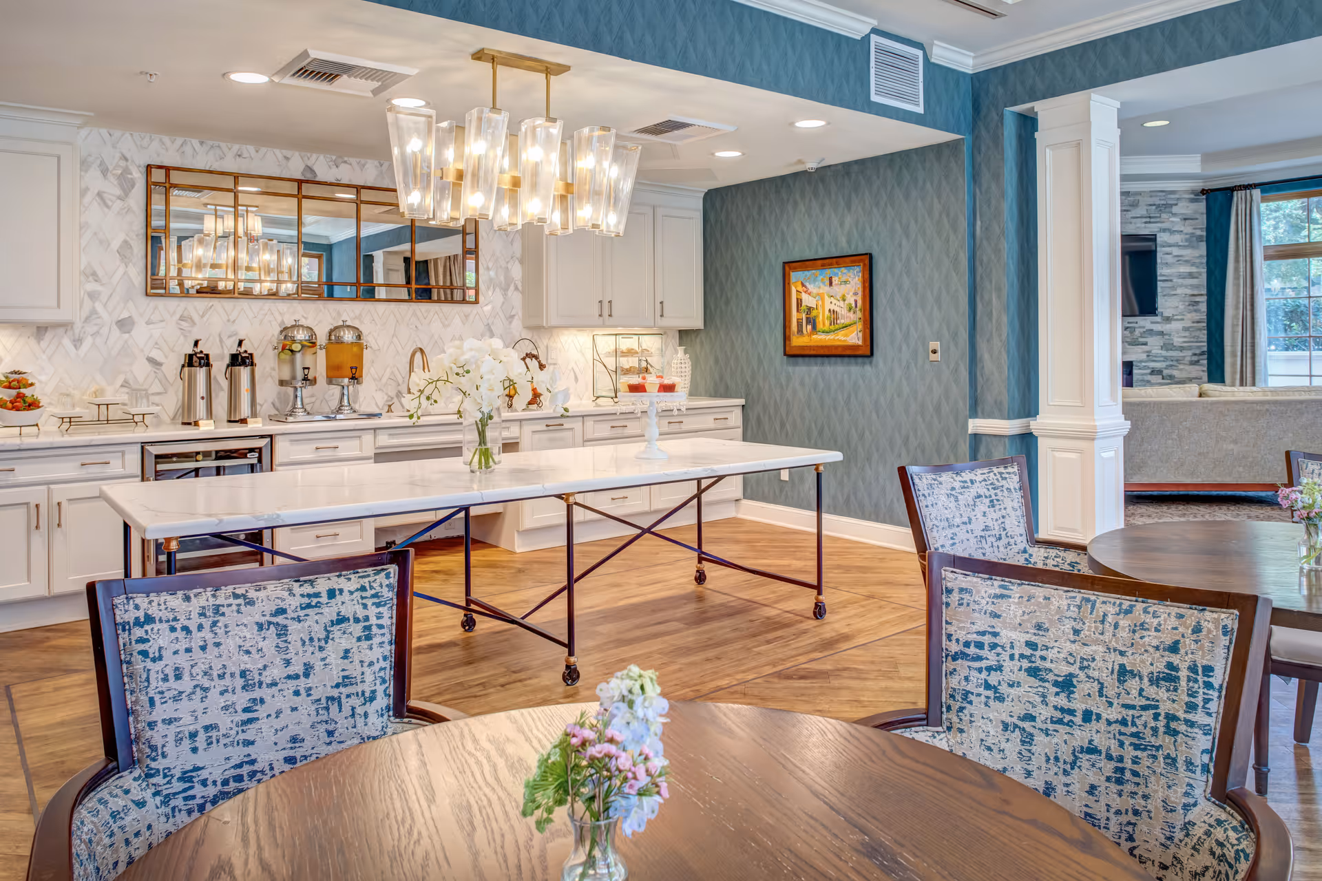 Bright communal dining and serving area with round tables, upholstered chairs, a long white island, and a chandelier.