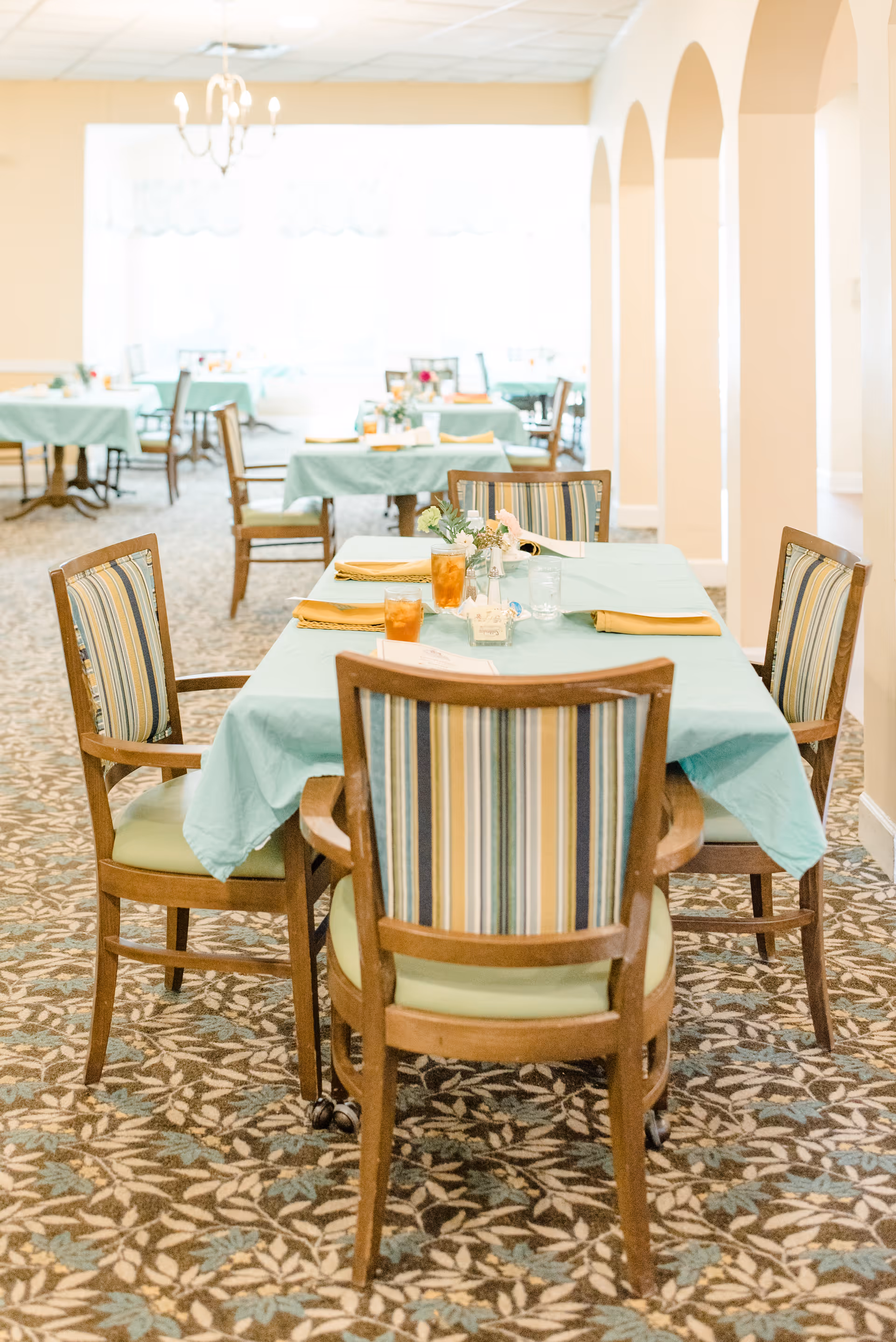 A bright dining room with tables covered in light blue tablecloths and set with yellow napkins, glasses of iced tea, and small floral centerpieces. The room features patterned carpet flooring, wooden chairs with striped upholstery, and arched openings along one side. A chandelier hangs from the ceiling near a large window letting in natural light.