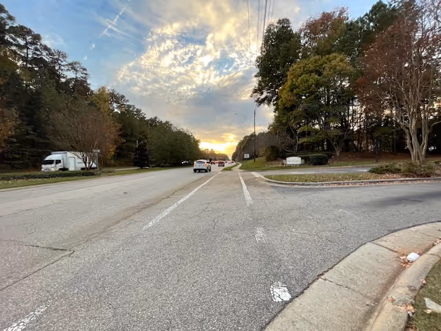 Wide two-lane road at sunset with cars, trees on both sides and the entrance sign to Brookdale Cary visible on the right.