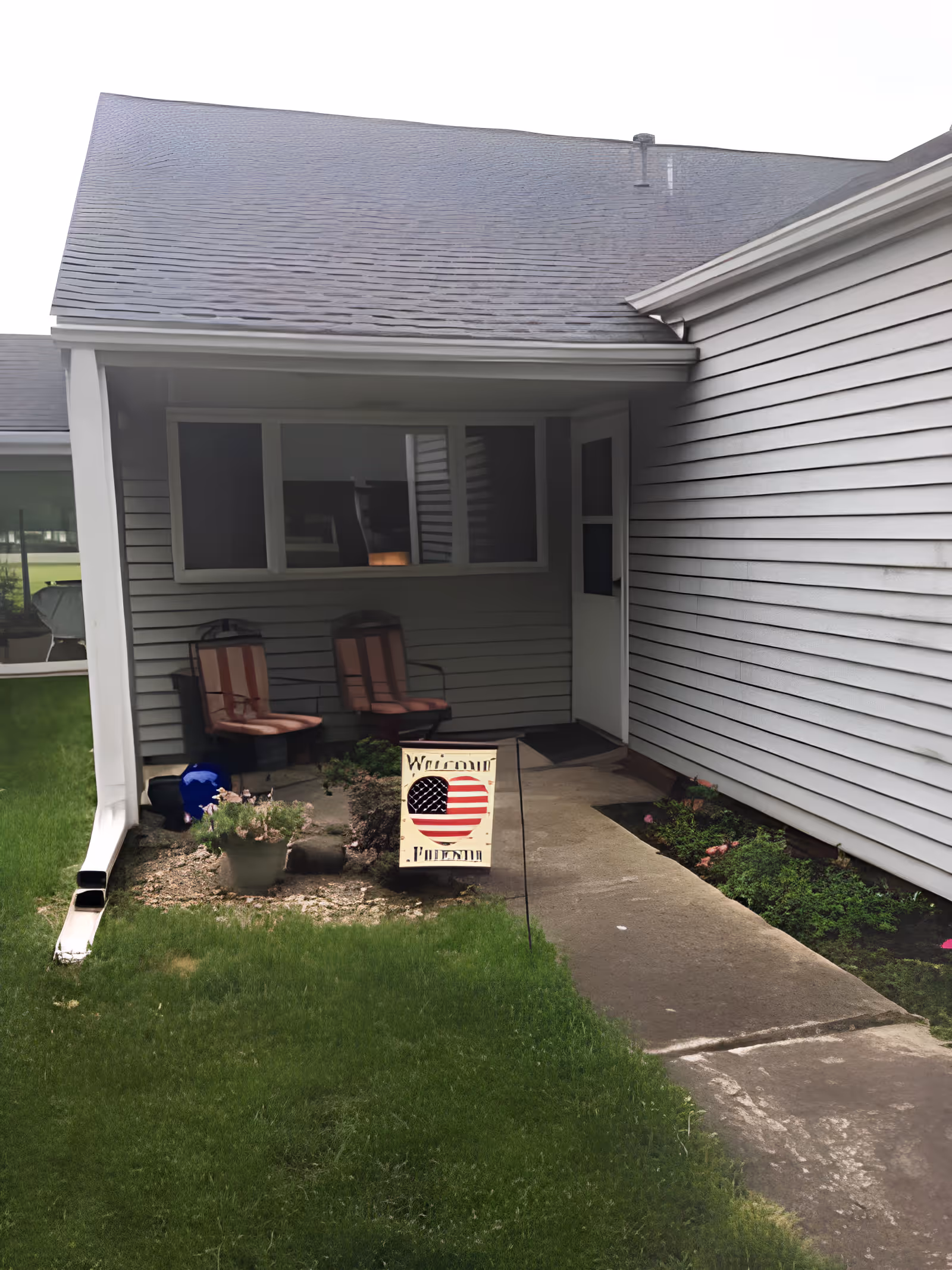 Covered porch area with two cushioned chairs, a small garden with plants, and a sign featuring an American flag heart and the word 'Welcome'. The porch is attached to a building with gray siding and a concrete walkway leading to a door.