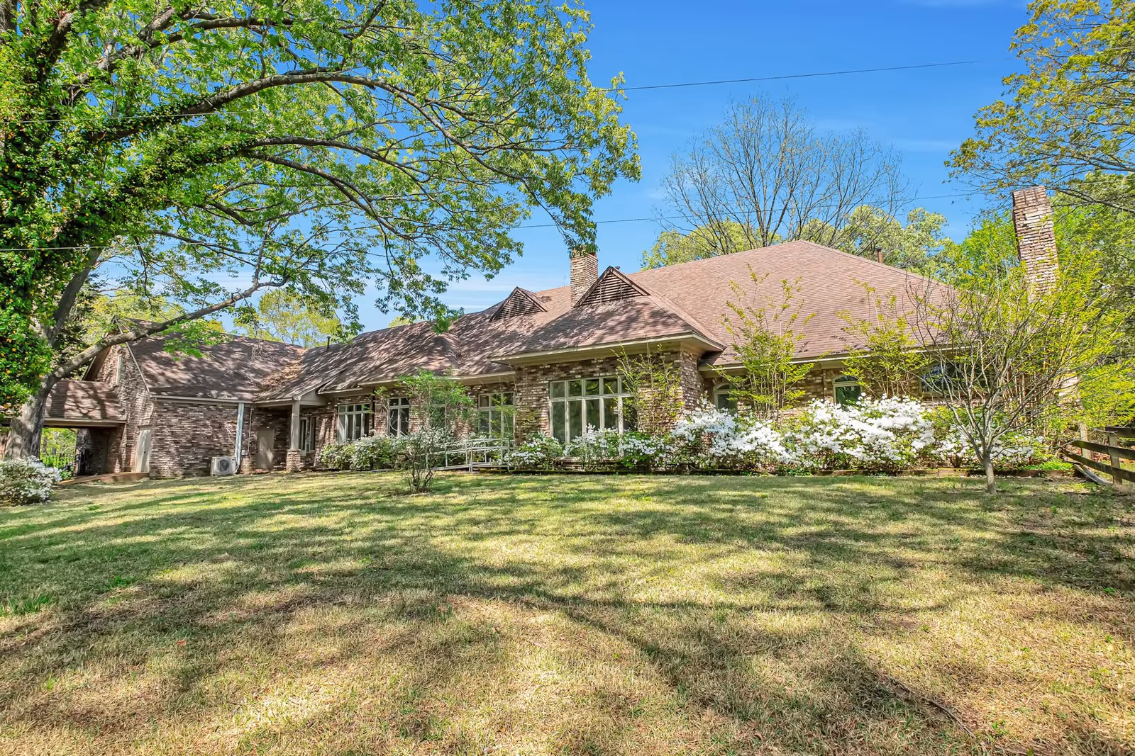 A large brick building with a brown roof surrounded by green trees and bushes with white flowers, under a clear blue sky. The building is set back from a grassy lawn with shadows from the trees.