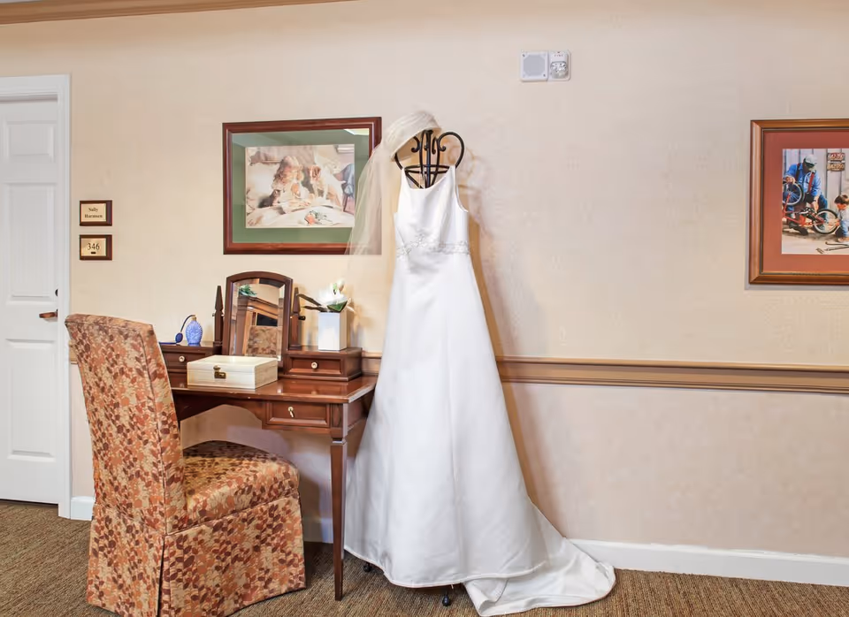 A white wedding dress on a mannequin hangs beside a wooden vanity and patterned chair in a warmly decorated interior room.
