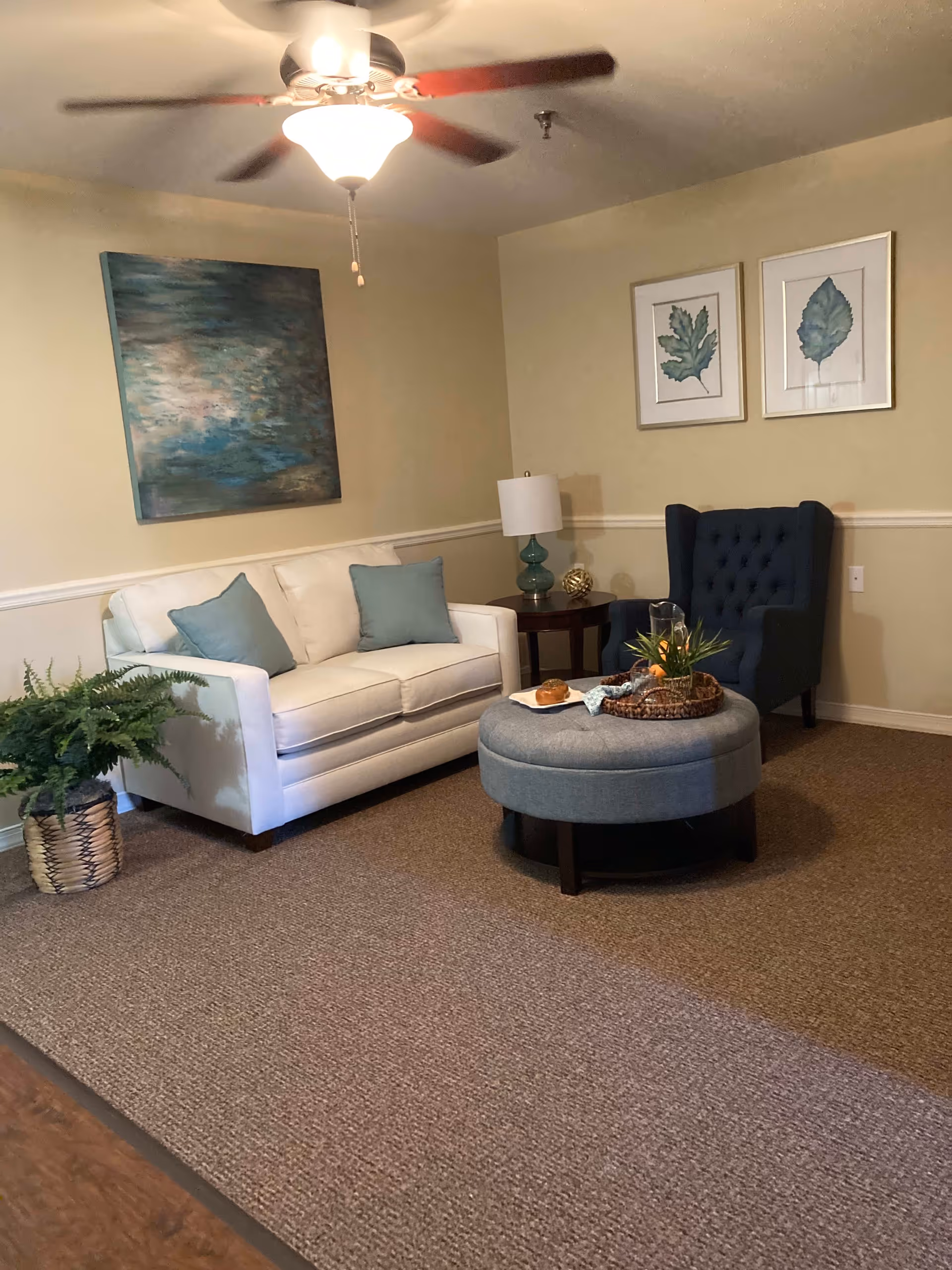 A cozy living room with a white sofa adorned with two light blue pillows, a dark blue tufted armchair, and a round gray ottoman with a tray holding a plant and a pastry. The walls are beige with white trim, decorated with two framed leaf prints and a large abstract painting. A ceiling fan with a light is on the ceiling, and a green potted fern sits on the floor beside the sofa.