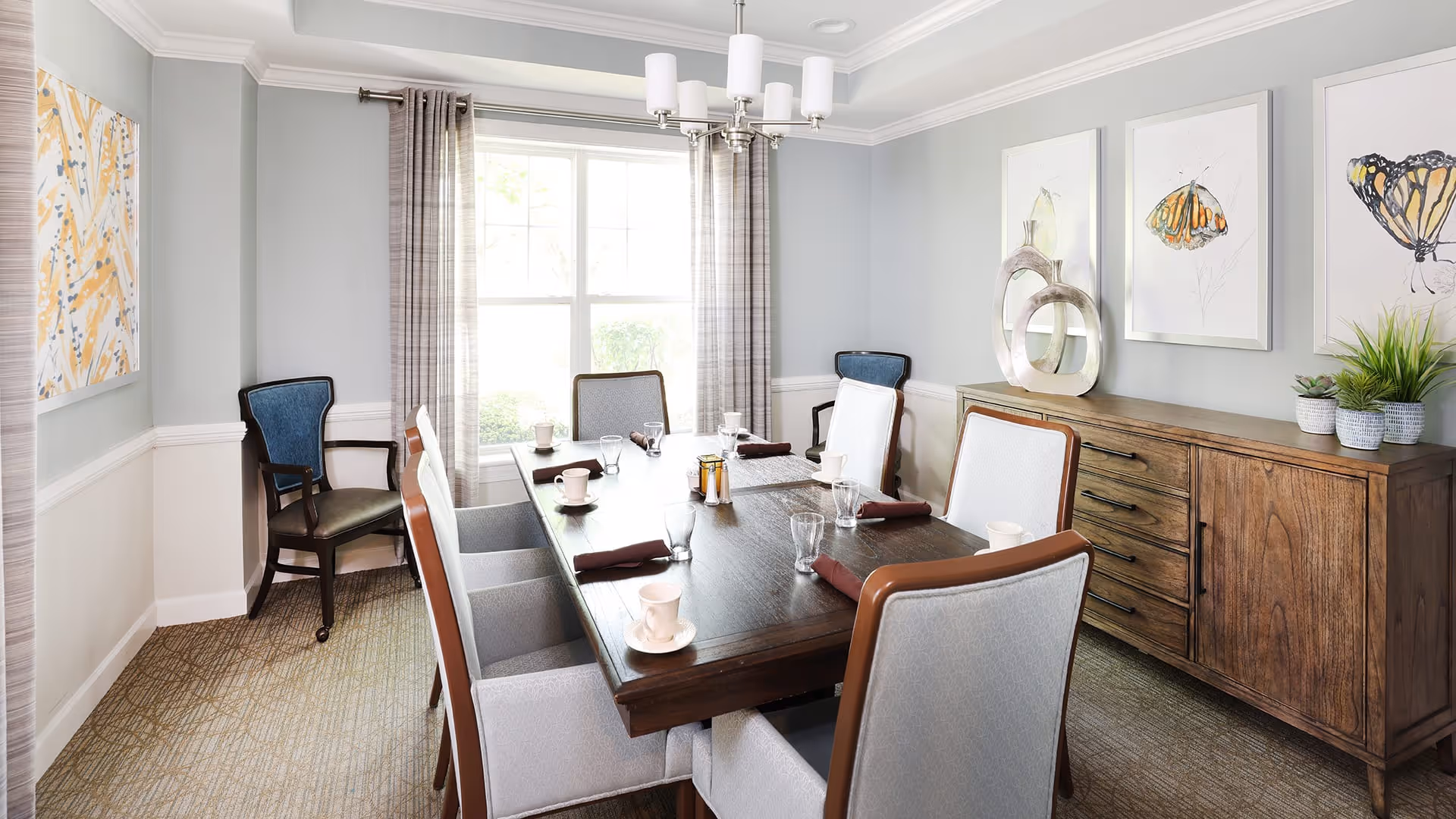 A bright dining room with a large wooden table set for six people. The table has cups, glasses, and folded napkins. There are six upholstered chairs around the table, with two additional wooden chairs with blue cushions against the walls. The room has light gray walls, a large window with plaid curtains, a wooden sideboard with decorative items and plants, and three framed butterfly artworks on the wall.