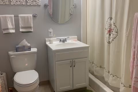 A clean bathroom with a white toilet, a white vanity with a sink, a round mirror above the sink, a tissue box on the toilet tank, two white towels hanging on a towel rack, and a shower curtain with a floral design.