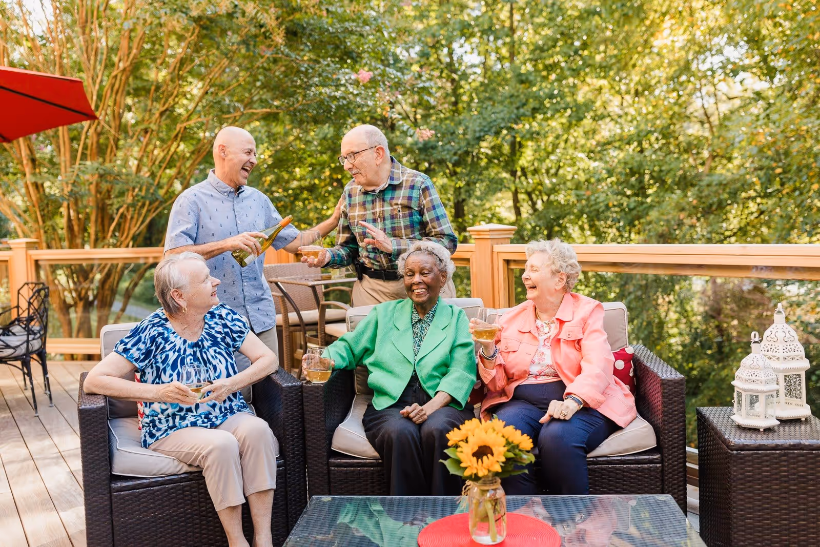 Five elderly people enjoying a social gathering on an outdoor patio with wicker furniture. Three women are seated on a cushioned sofa, holding drinks and smiling, while two men stand behind them, one pouring a drink. The background shows green trees and a wooden railing, with decorative lanterns and a vase with sunflowers on the table.