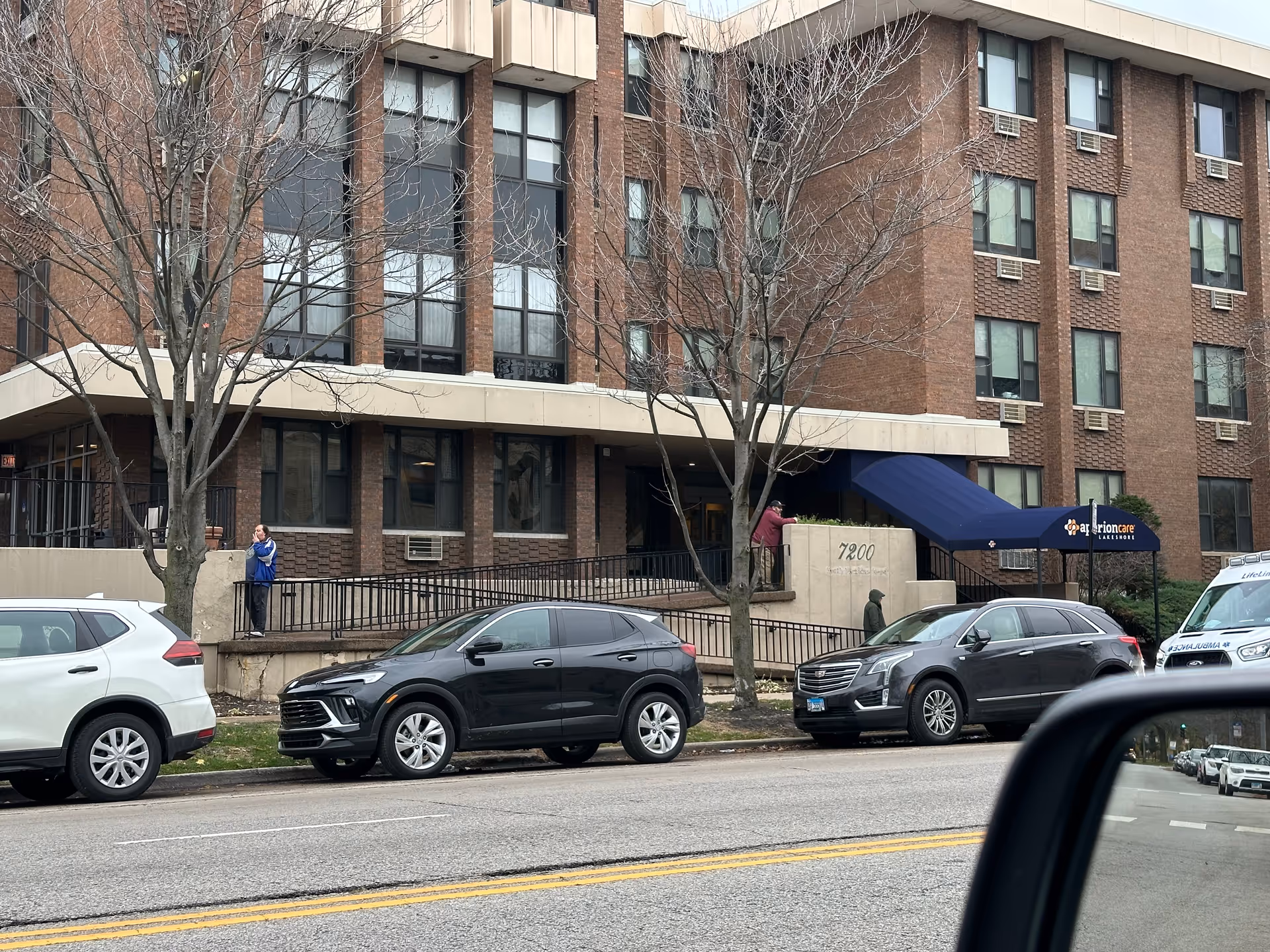 Exterior view of a multi-story brick building with several windows and air conditioning units. There is a blue awning over the entrance with the text 'Aperion Care Lakeshore'. Several cars are parked along the street in front of the building, and a few people are visible near the entrance and sidewalk. Leafless trees are also present in front of the building.