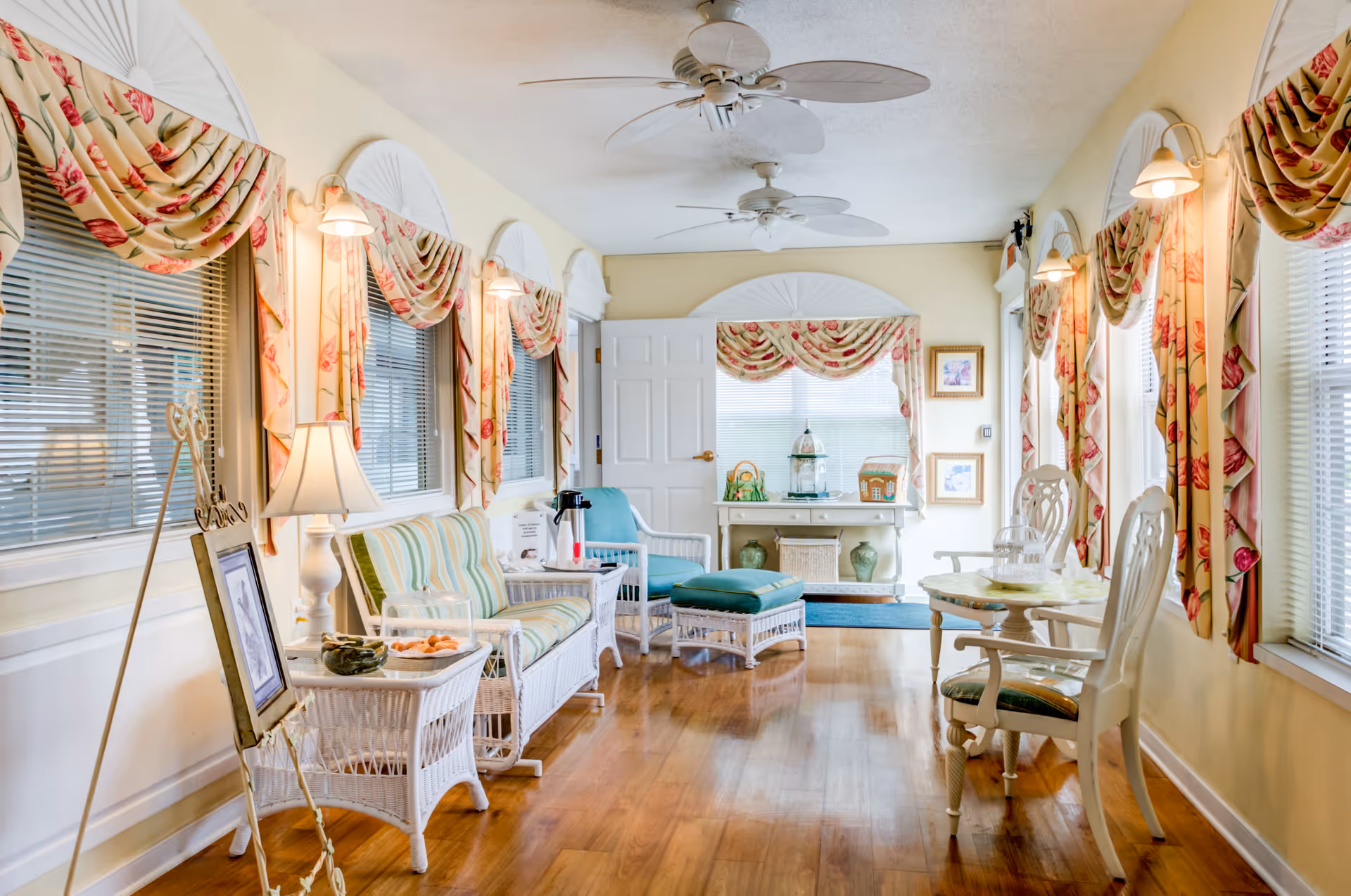 A bright and cozy living room area with wooden floors, white wicker furniture including a loveseat, chairs, and a small table. The room has multiple windows adorned with floral-patterned curtains and valances. Ceiling fans with leaf-shaped blades hang from the ceiling, and there are wall-mounted lamps providing warm lighting. A small table with two chairs is positioned near the windows on the right side, and a console table with decorative items is at the far end of the room.