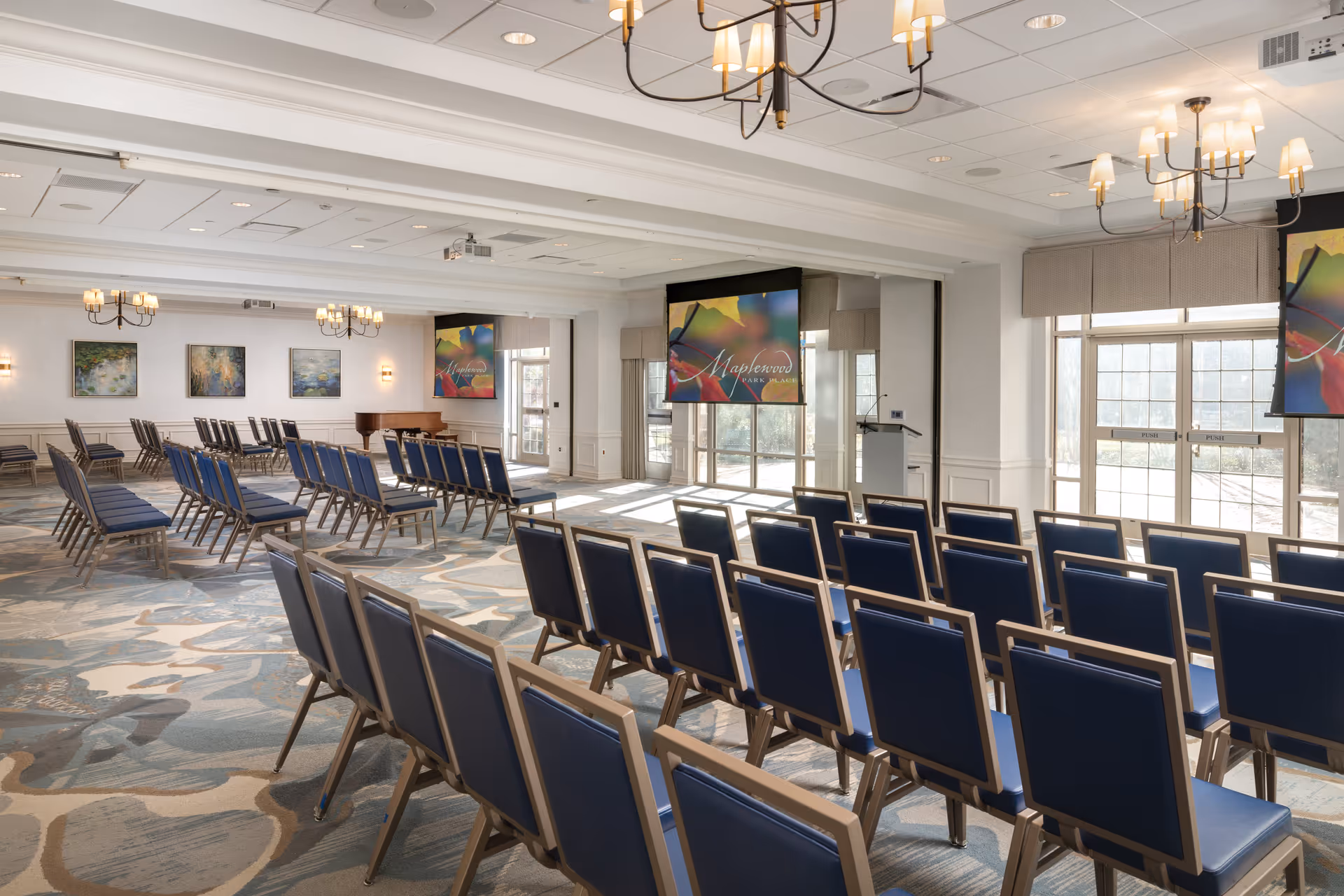 A spacious conference or meeting room with rows of blue cushioned chairs arranged facing two large projection screens displaying the Maplewood Park Place logo. The room has large windows letting in natural light, chandeliers hanging from the ceiling, and framed artwork on the walls.