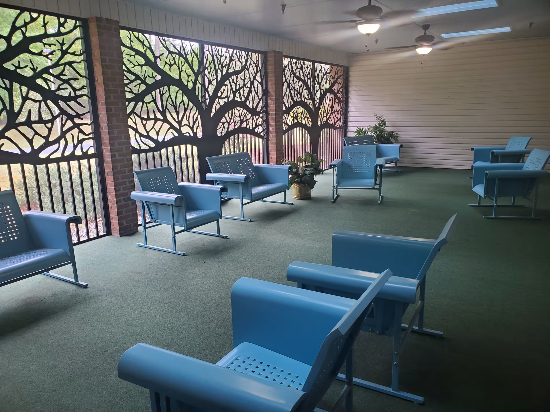 A covered outdoor seating area with multiple light blue metal chairs arranged along the walls and in the center. The area has green carpet flooring and decorative metal panels with tree branch designs on the railing. Ceiling fans with lights are mounted on the white ceiling, and there are a few potted plants placed near the walls.
