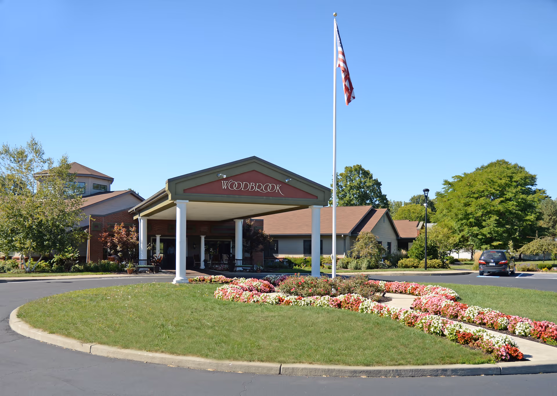 Front exterior view of Woodbrook Assisted Living Residence, Inc. building with a covered entrance, an American flag on a flagpole, a circular driveway, and landscaped flower beds with green trees and a clear blue sky in the background.