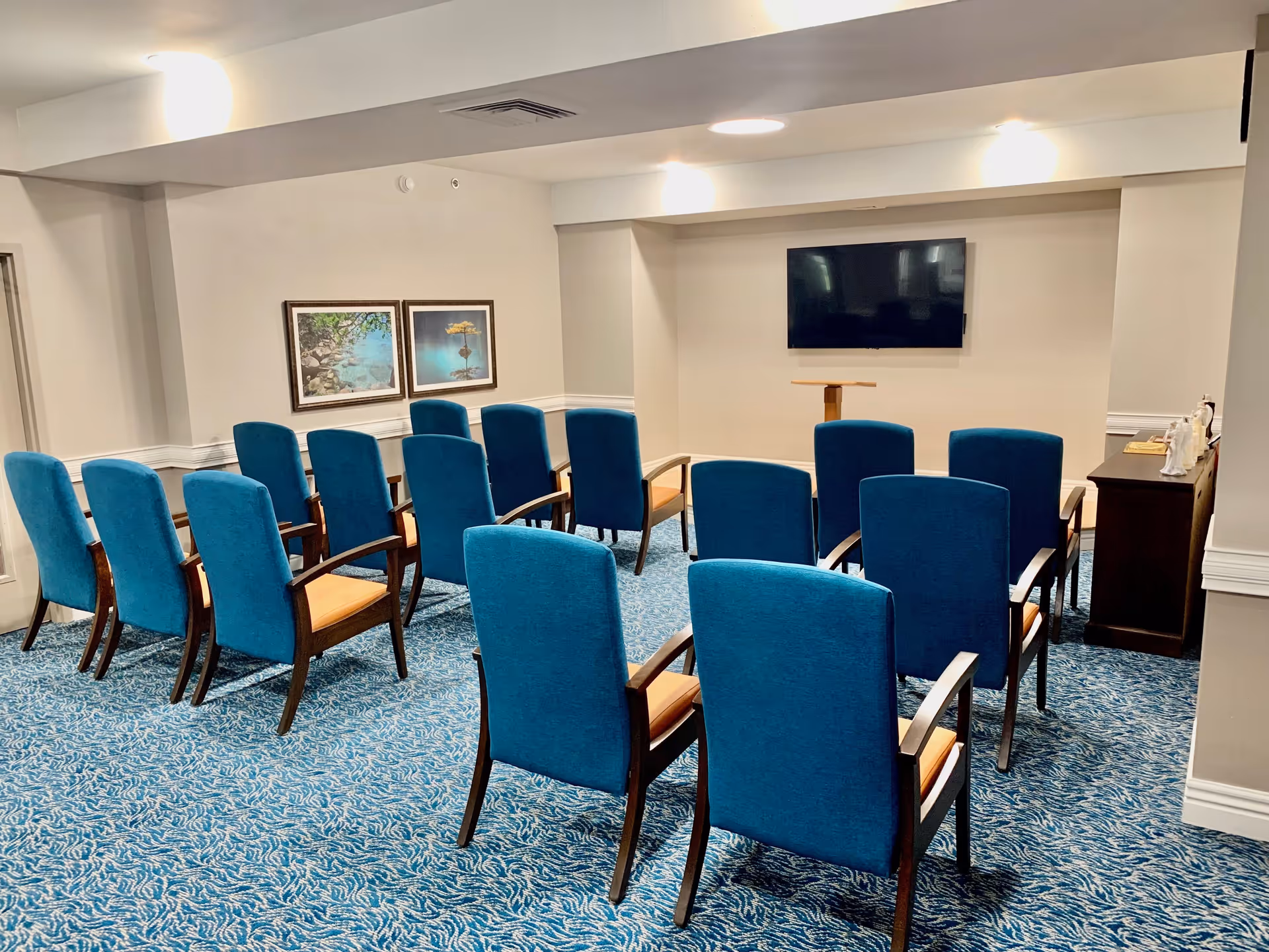 A small meeting or presentation room with two rows of blue upholstered chairs facing a wall-mounted flat screen TV. The room has blue patterned carpet, light-colored walls, two framed pictures on one wall, and a wooden podium in front of the TV. There is also a dark wooden cabinet with decorative items on the right side of the room.