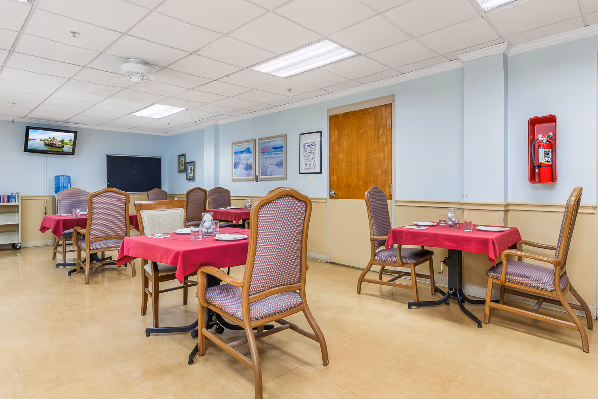 A dining room with several tables covered in red tablecloths, each set with glasses, plates, and napkins. The room has wooden chairs with cushioned seats and backs, light blue walls with framed pictures, a wooden door, a fire extinguisher mounted on the wall, and a wall-mounted TV displaying a scenic image.