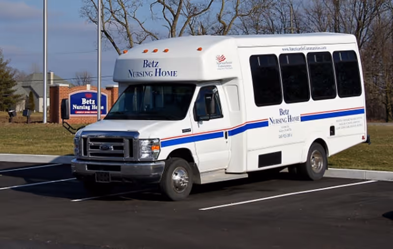 A white shuttle bus parked in a parking lot with 'Betz Nursing Home' written on the side and front. In the background, there is a sign for Betz Nursing Home and some trees without leaves.