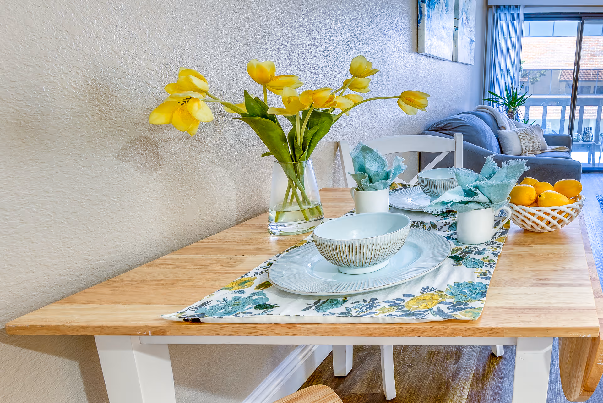 A wooden dining table set with two place settings including bowls and plates on a floral table runner. A clear vase with yellow tulips and two small white mugs with teal cloth napkins are on the table. A basket of lemons is also present. In the background, there is a gray sofa with pillows and a large window with vertical blinds letting in natural light.