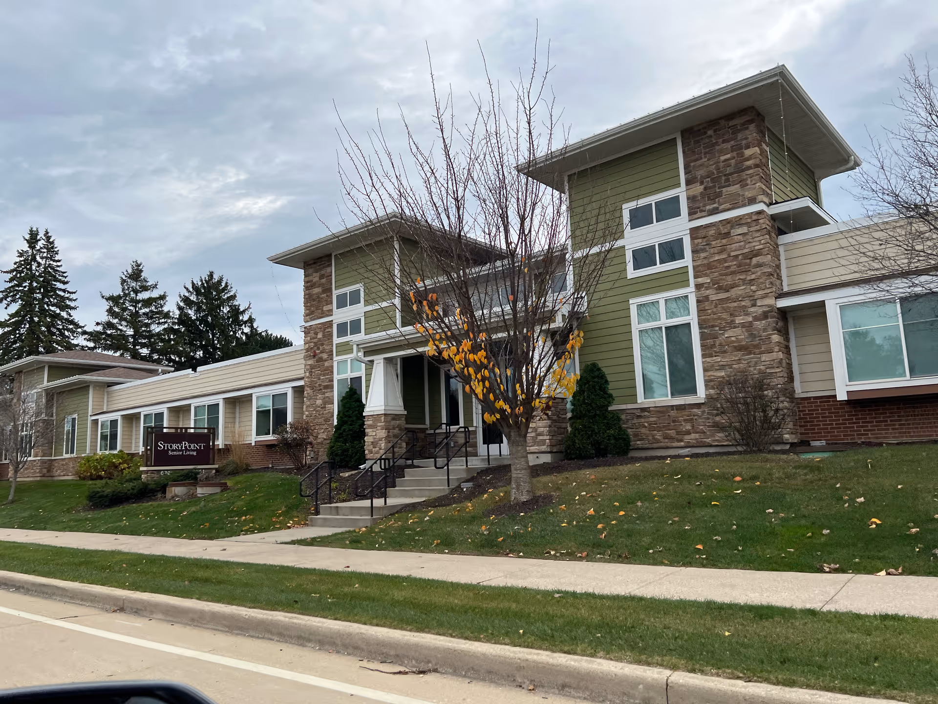 Exterior view of StoryPoint Mequon senior living facility showing a modern building with green and stone facade, large windows, and a small tree with autumn leaves in front. There is a sidewalk and a grassy area leading up to the entrance.