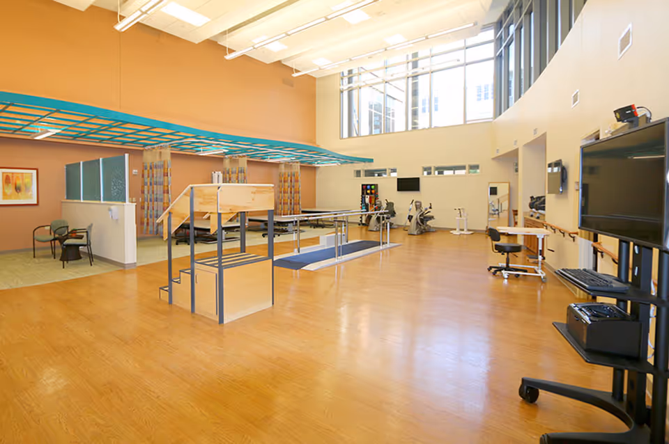 Bright, open rehabilitation/therapy room with parallel bars, exercise equipment, therapy stairs and seating under large windows.